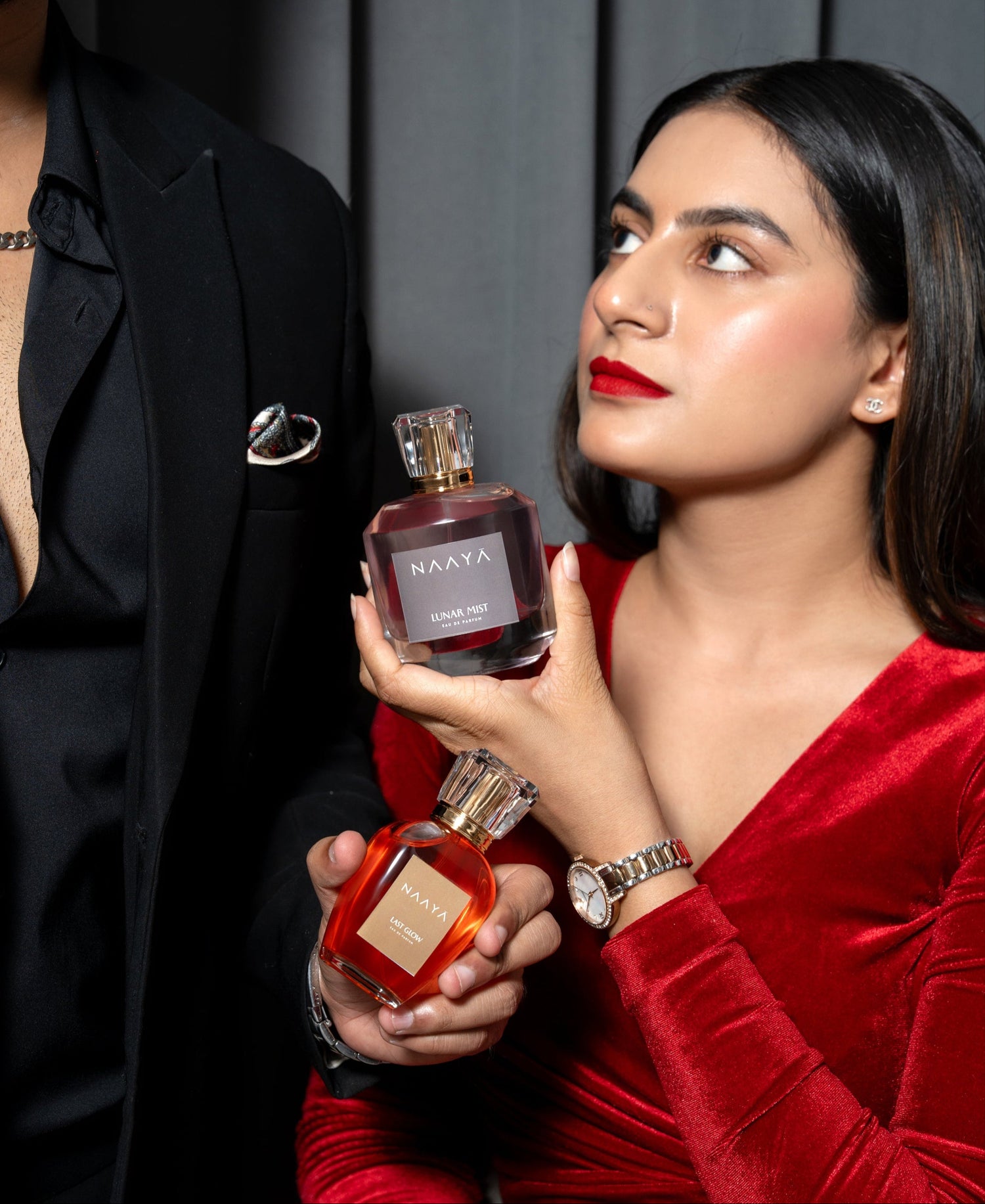 Woman in a red dress holding a perfume bottle with a man in a black suit next to her.